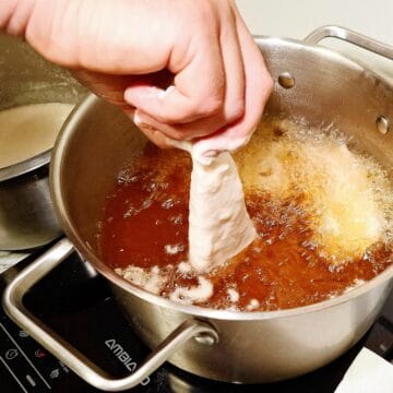 A pot with hot oil frying fish pieces and one piece of fish is half dipped into the oil. A stainless steel bowl next to it with tempura batter