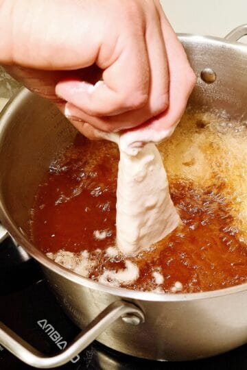 A pot with hot oil frying fish pieces and one piece of fish is half dipped into the oil. A stainless steel bowl next to it with tempura batter