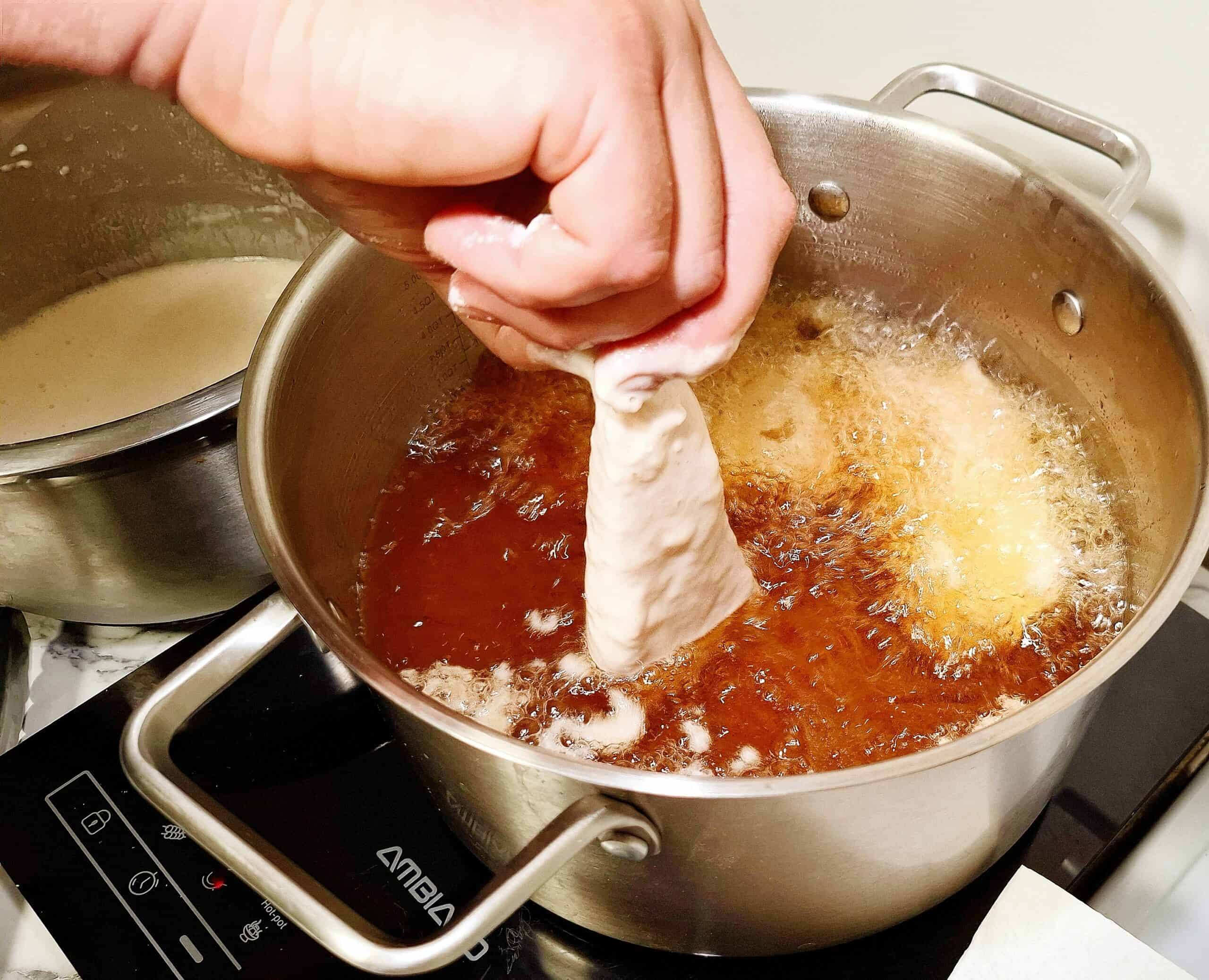 A pot with hot oil frying fish pieces and one piece of fish is half dipped into the oil. A stainless steel bowl next to it with tempura batter