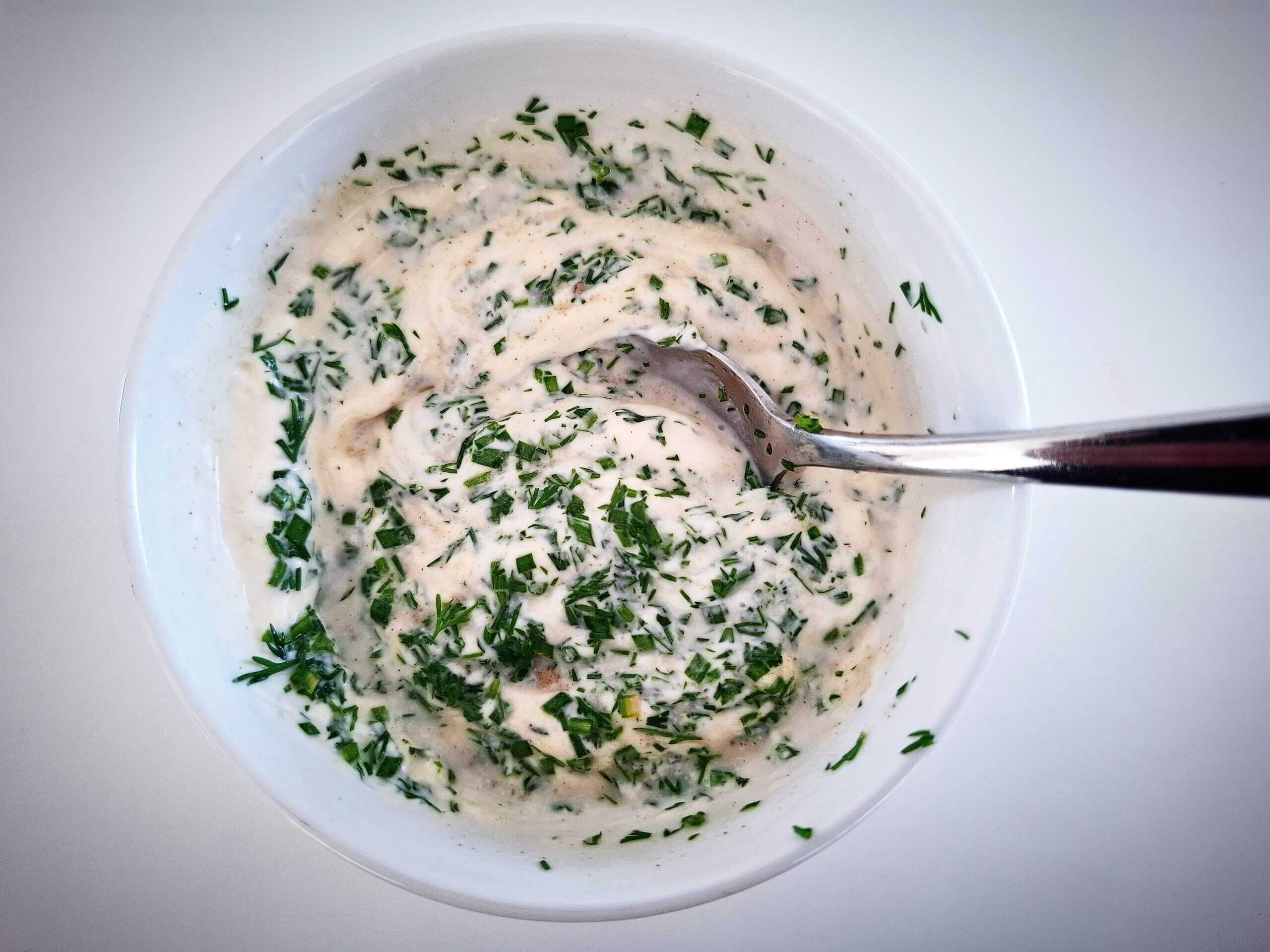 mixing the skagen dressing in a white bowl with a silver spoon on a white table.The fresh herbs are on top and not fully mixed in.