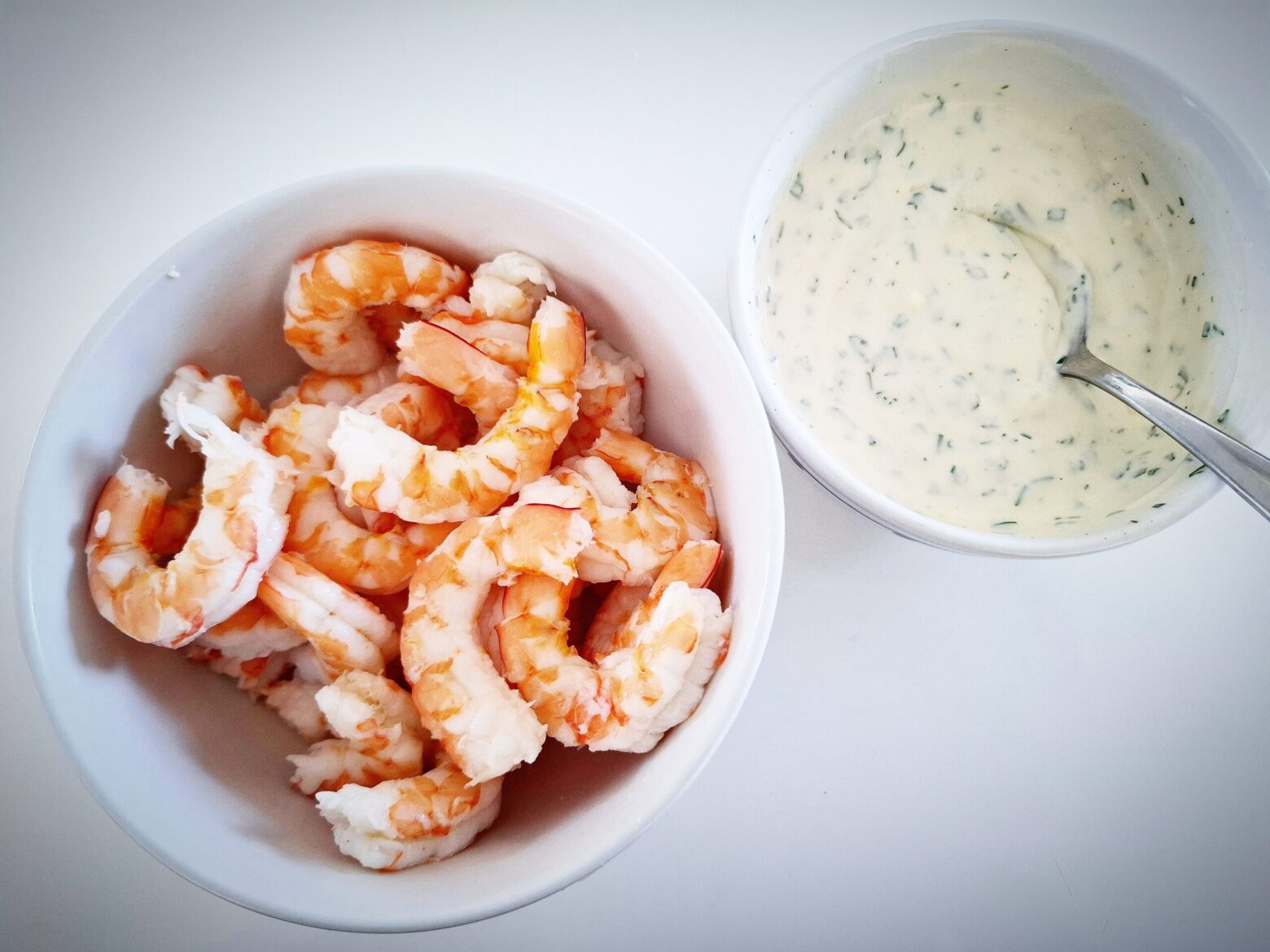 Peeled prawns in a white bowl, and skagen dressing in another white bowl with a silver spoon in it, on a white table.