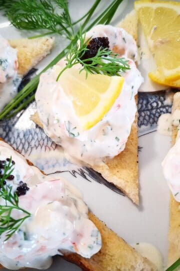 Four triangular pieces of skagen toast on a platter topped with black lumpfish caviar and garnished with a small slice of lemon and fresh dill. Fresh chives, dill leaves and lemon slices places around the skagen toast on the platter.
