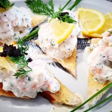 Four triangular pieces of skagen toast on a platter topped with black lumpfish caviar and garnished with a small slice of lemon and fresh dill. Fresh chives, dill leaves and lemon slices places around the skagen toast on the platter.