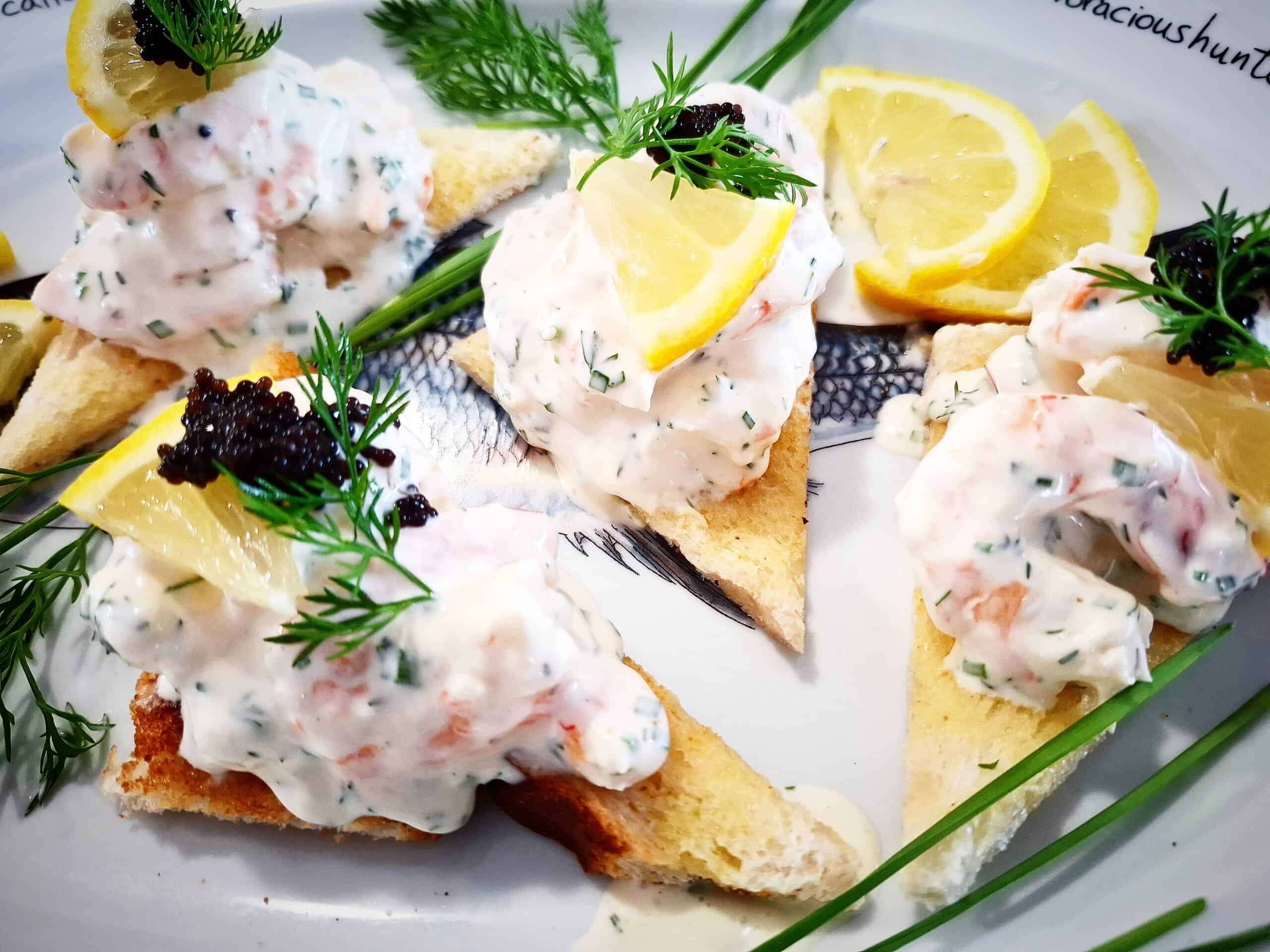 Four triangular pieces of skagen toast on a platter topped with black lumpfish caviar and garnished with a small slice of lemon and fresh dill. Fresh chives, dill leaves and lemon slices places around the skagen toast on the platter.