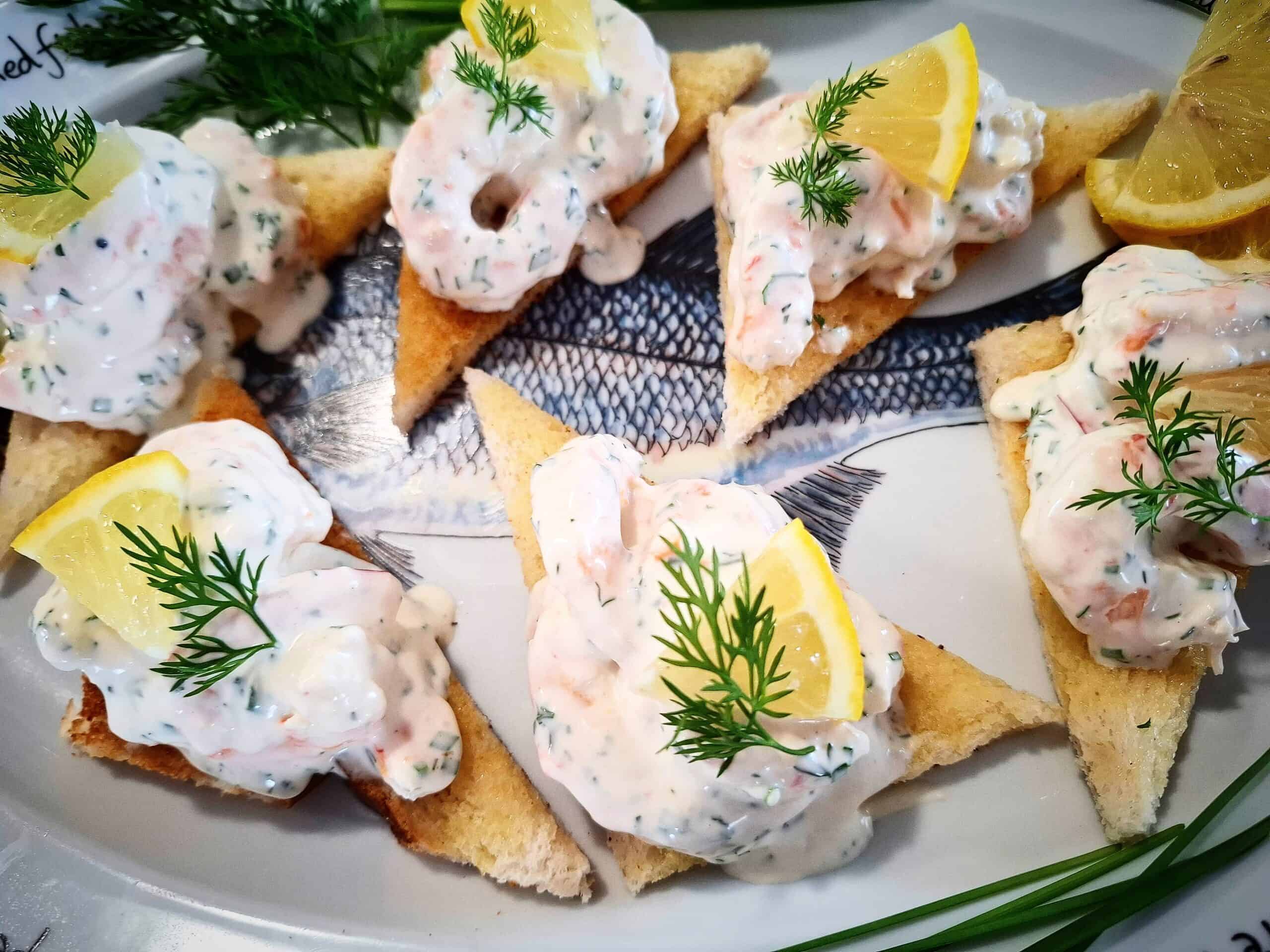 Six triangular pieces of toast on a platter topped with skagen and garnished with a small slice of lemon and fresh dill. Fresh chives, dill leaves and lemon slices places around the toast on the platter.