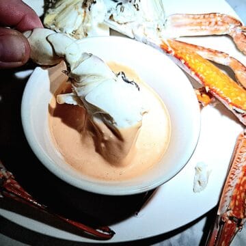 Seafood cocktail sauce in a round ramekin on a white plate with crab pieces on it. A chunk of crab is dipping into the sauce.