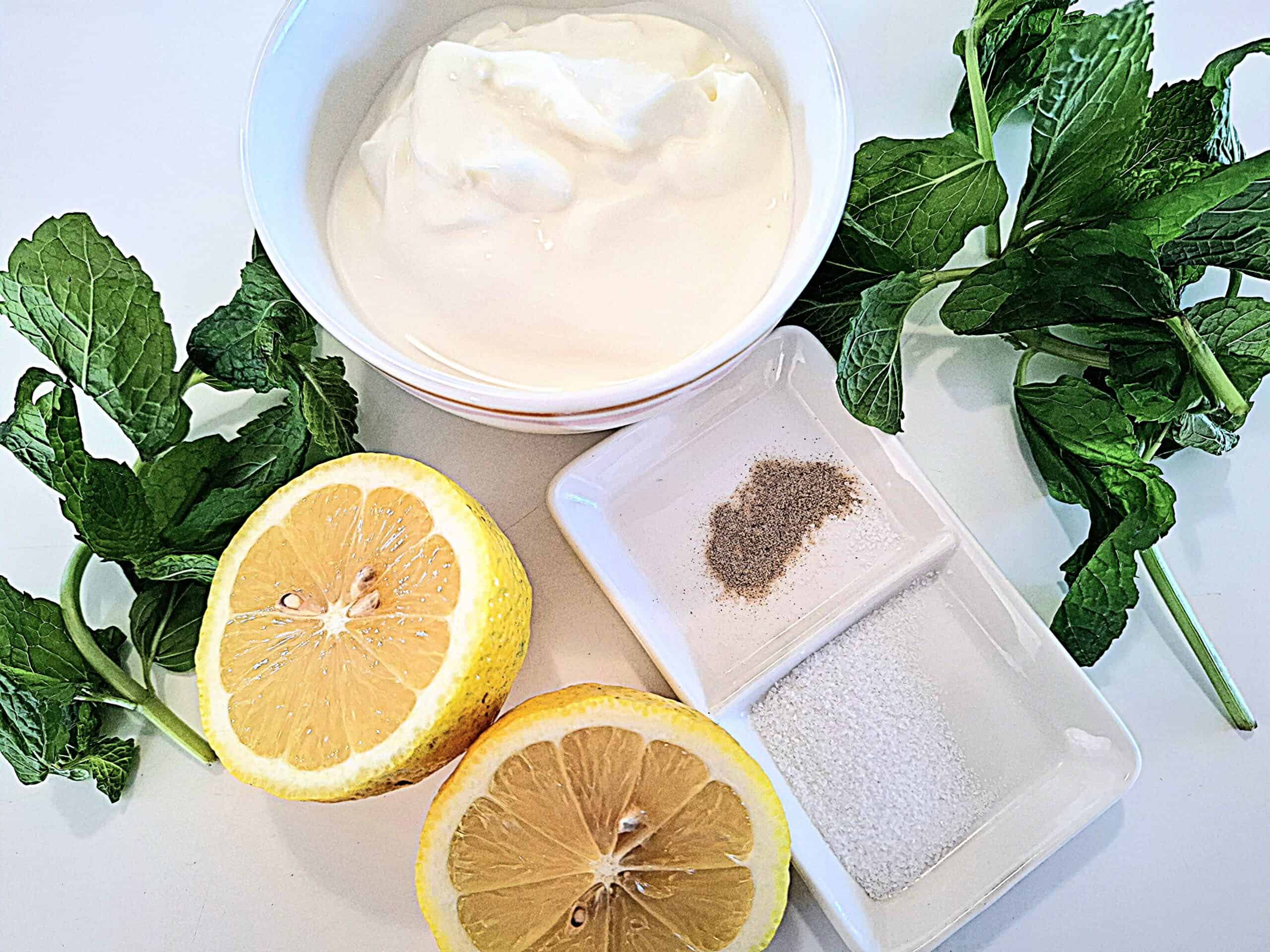 Ingredient for mint and yoghurt sauce. A white bowl of yoghurt, a double ramekin with salt and pepper, a lemon cut in half and fresh mint leaves on a white table.