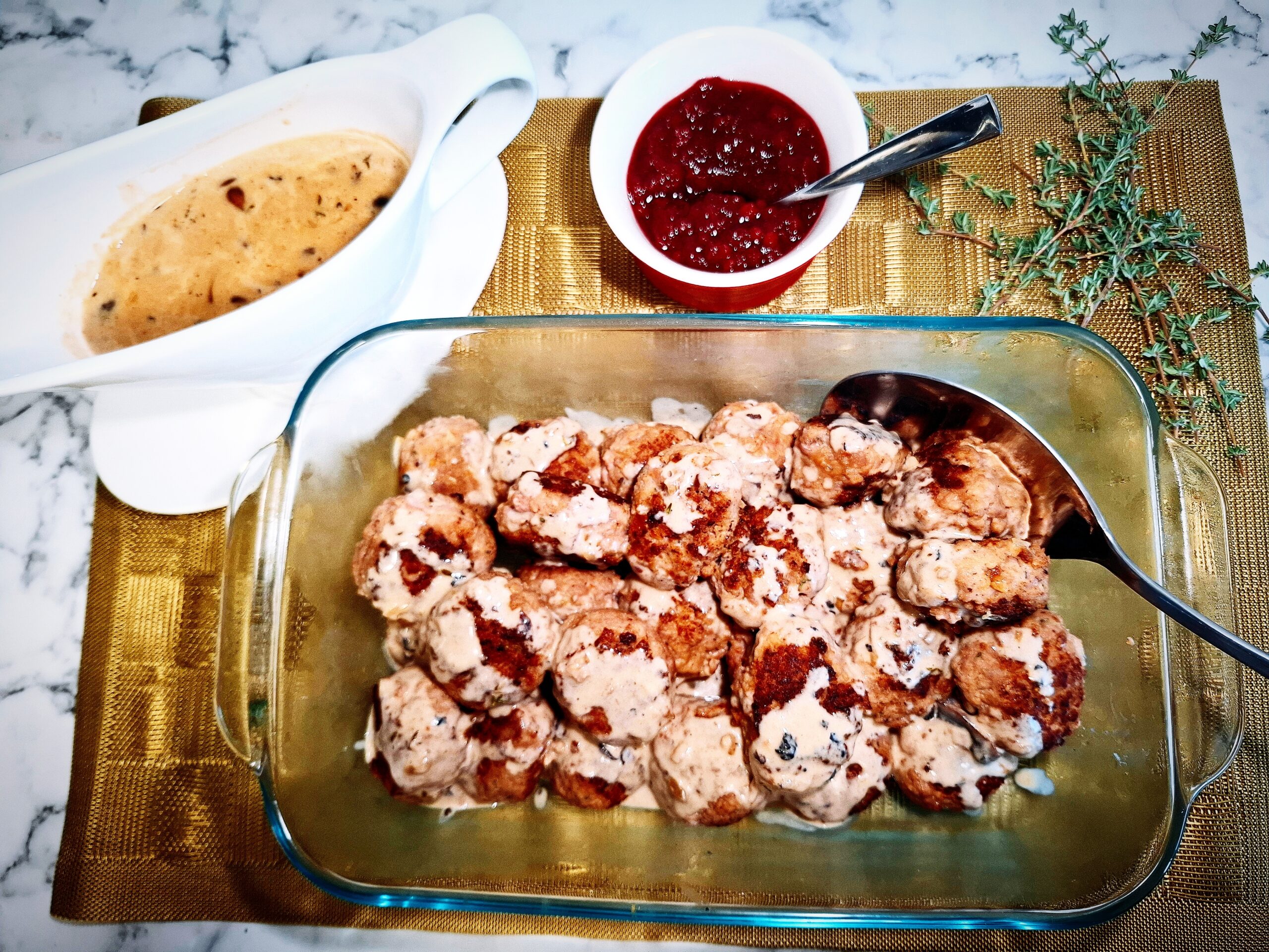 Meatballs drizzled with creamy mushroom sauce in a glass oven proof dish with a serving spoon in it. A white sauce pot filled with creamy mushroom sauce, a red and white ramekin with lingon berry jam and some fresh thyme sprigs on a golden placemat