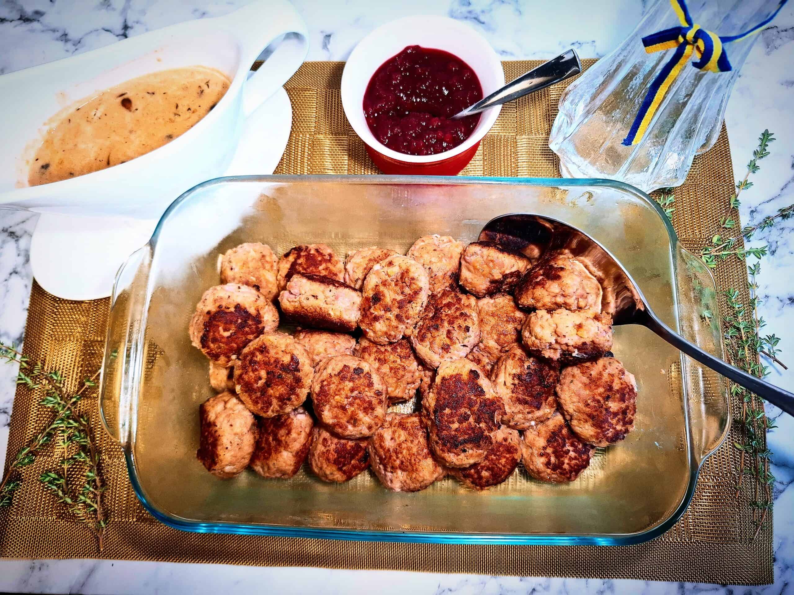 Meatballs in a glass oven proof dish with a serving spoon in it. A white sauce pot filled with creamy mushroom sauce, a red and white ramekin with lingonberry jam, fresh thyme sprigs and a glass vase with blue and yellow ribbon on a golden place mat.