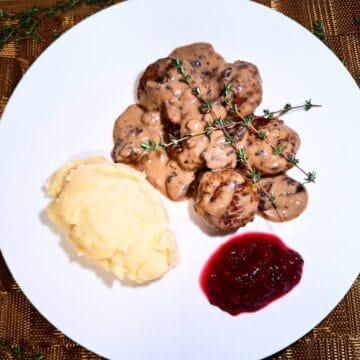 5 meatballs covered with a creamy mushroom sauce topped with thyme sprigs, mash potato and lingonberry jam on a white plate on a golden place mat
