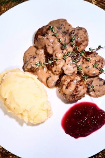 5 meatballs covered with a creamy mushroom sauce topped with thyme sprigs, mash potato and lingonberry jam on a white plate on a golden place mat