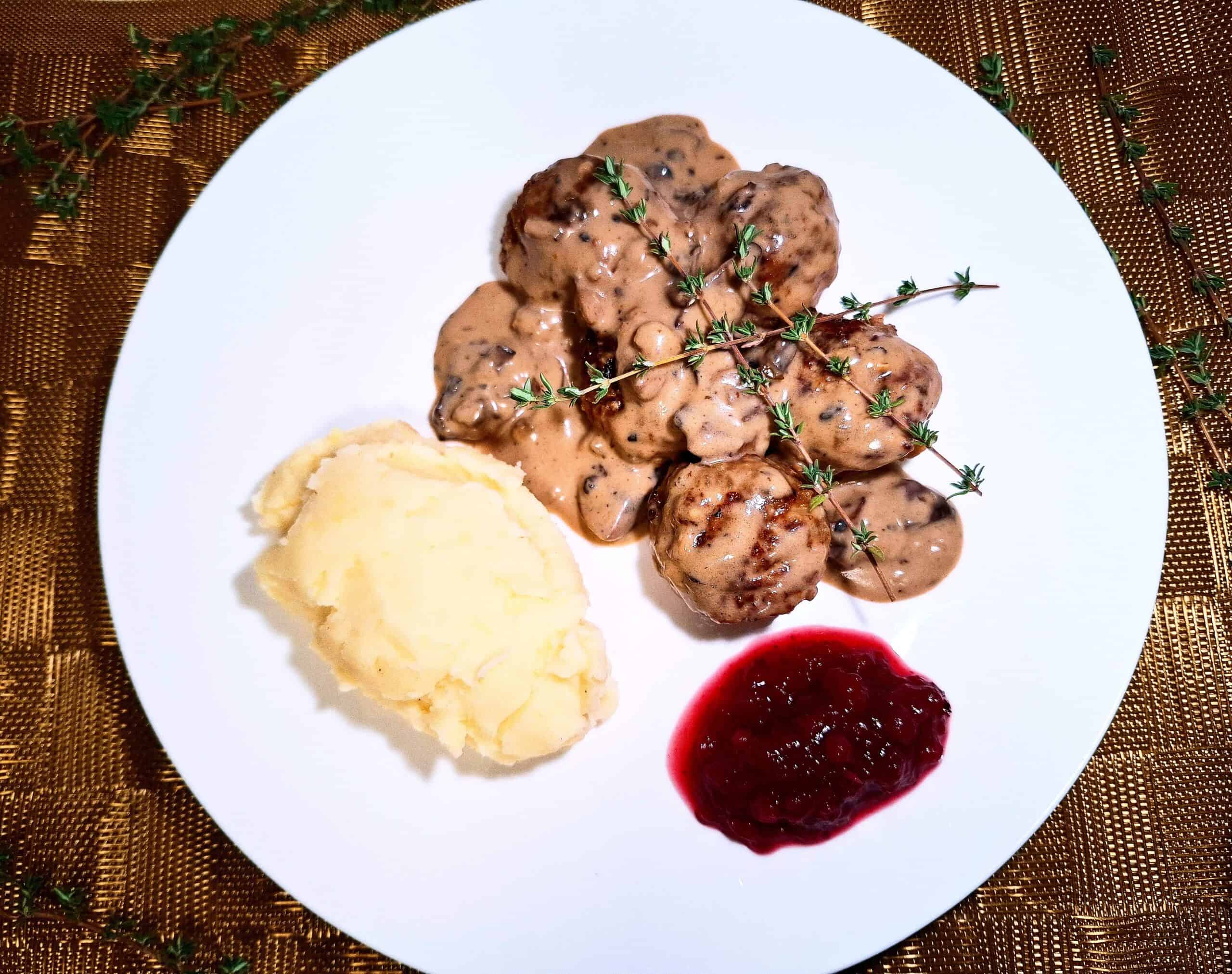 5 meatballs covered with a creamy mushroom sauce topped with thyme sprigs, mash potato and lingonberry jam on a white plate on a golden place mat