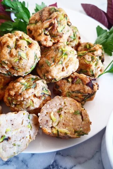 Cooked zucchini balls on a white plate, one cut in half, and next to it a white ramekin with sauce in it. Fresh herbs and lemons next to the plate.