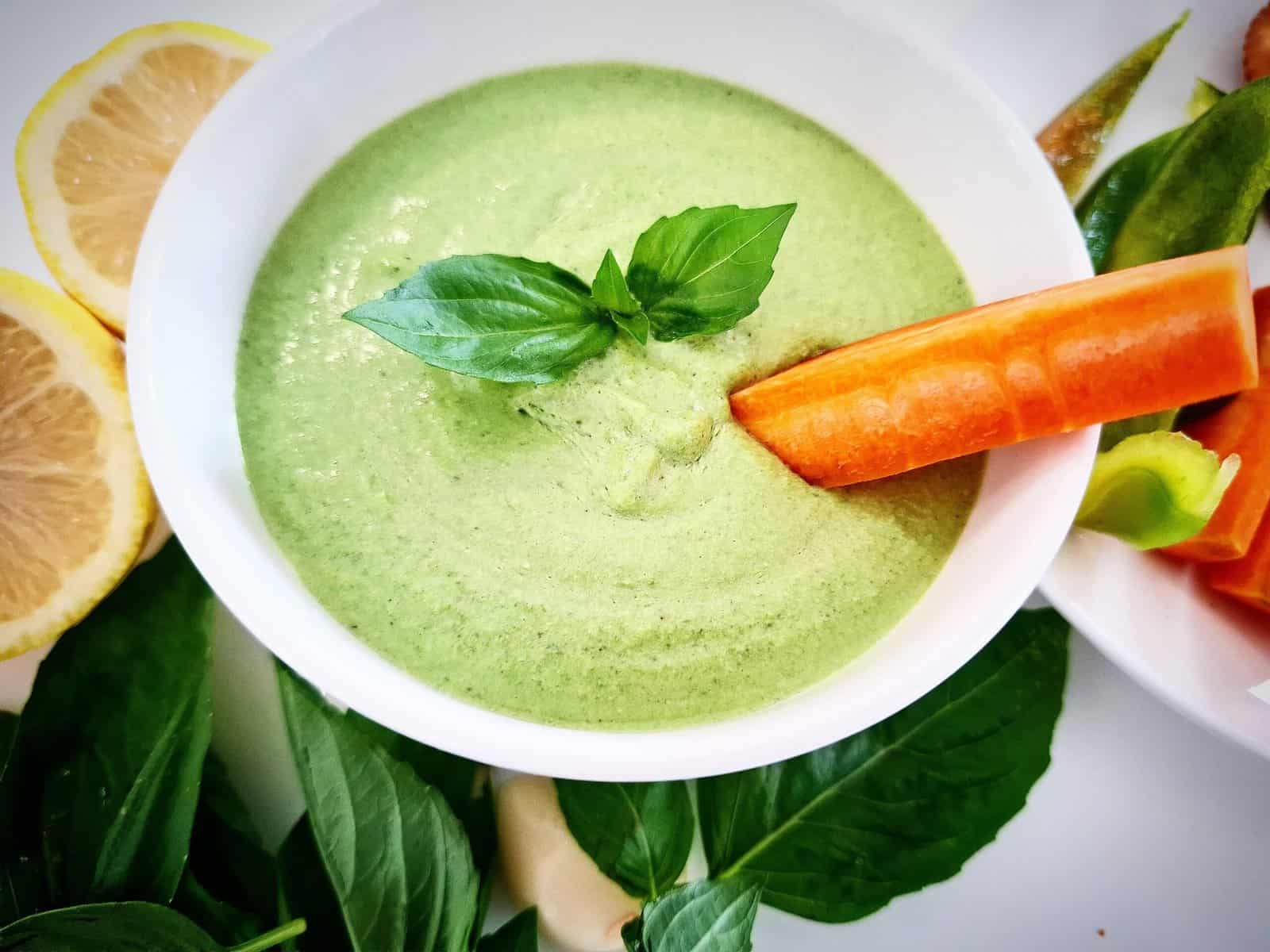 Basil dip in a white ramekin topped with basil leaves. Half a lemon a peeled garlic clove and fresh basil leaves next to it. A carrot stick being dipped into the basil dip.