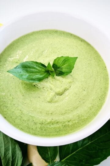 Basil dip in a white ramekin topped with fresh basil leaves. Half a lemon, a peeled garlic clove, fresh basil leaves and a plate with vegetable sticks next to it