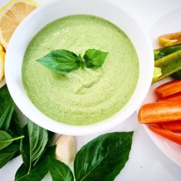 Basil dip in a white ramekin topped with basil leaves. Half a lemon, a peeled garlic clove, fresh basil leaves and a plate with vegetable sticks next to it