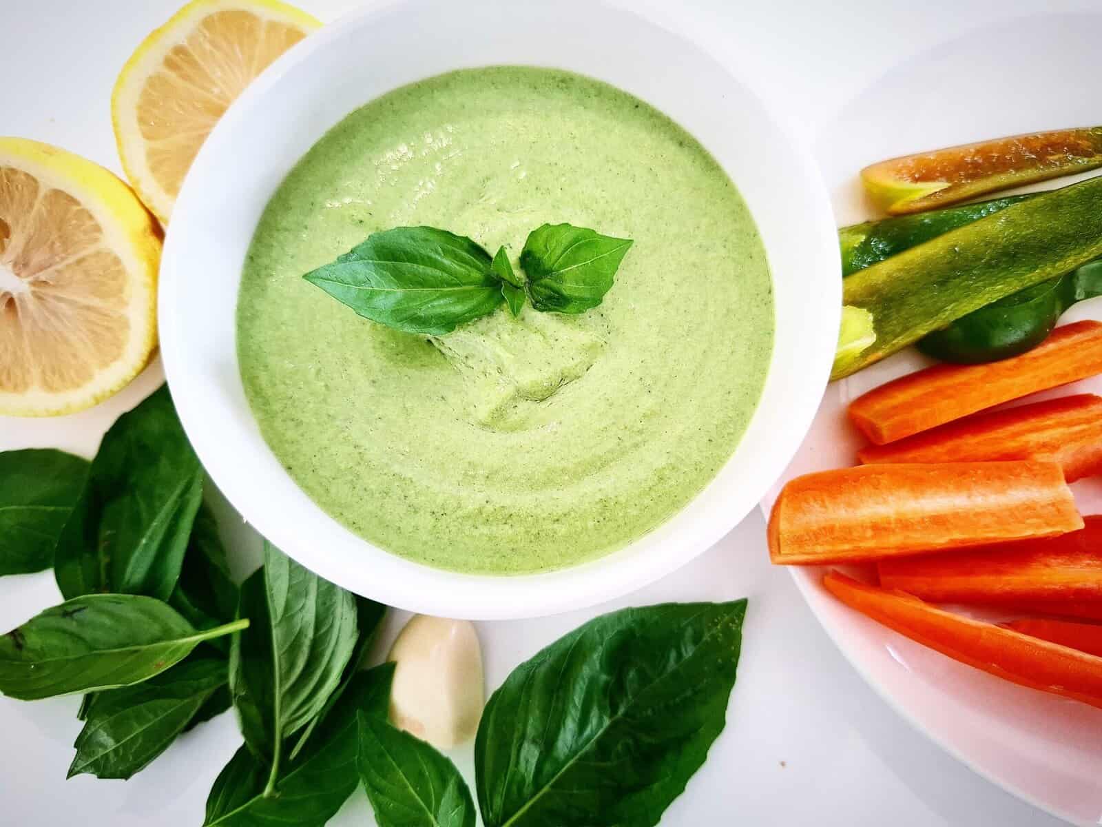 Basil dip in a white ramekin topped with basil leaves. Half a lemon, a peeled garlic clove, fresh basil leaves and a plate with vegetable sticks next to it