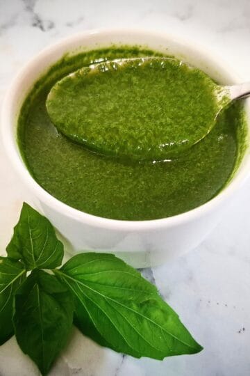 Basil garlic oil in a white ramekin with a spoon holding up some of the oil. Fresh basil leaves in front of it