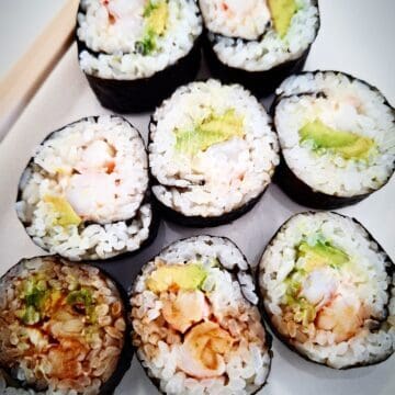 Eight pieces of prawn and avocado sushi on a white plate with two chop sticks next to them on the left hand side. Soy sauce drizzled on top of the front three pieces.