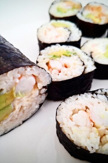 A hand roll and eight pieces of prawn and avocado sushi on a white plate. Soy sauce drizzled on top of the three pieces at the back .