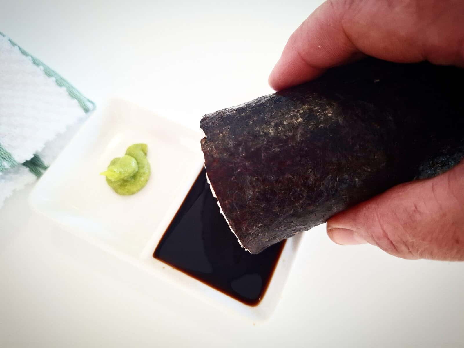 A hand holding a sushi hand roll with a double ramekin underneath with soy sauce and wasabi in it. A white towel with green stripes next to it on a white table.