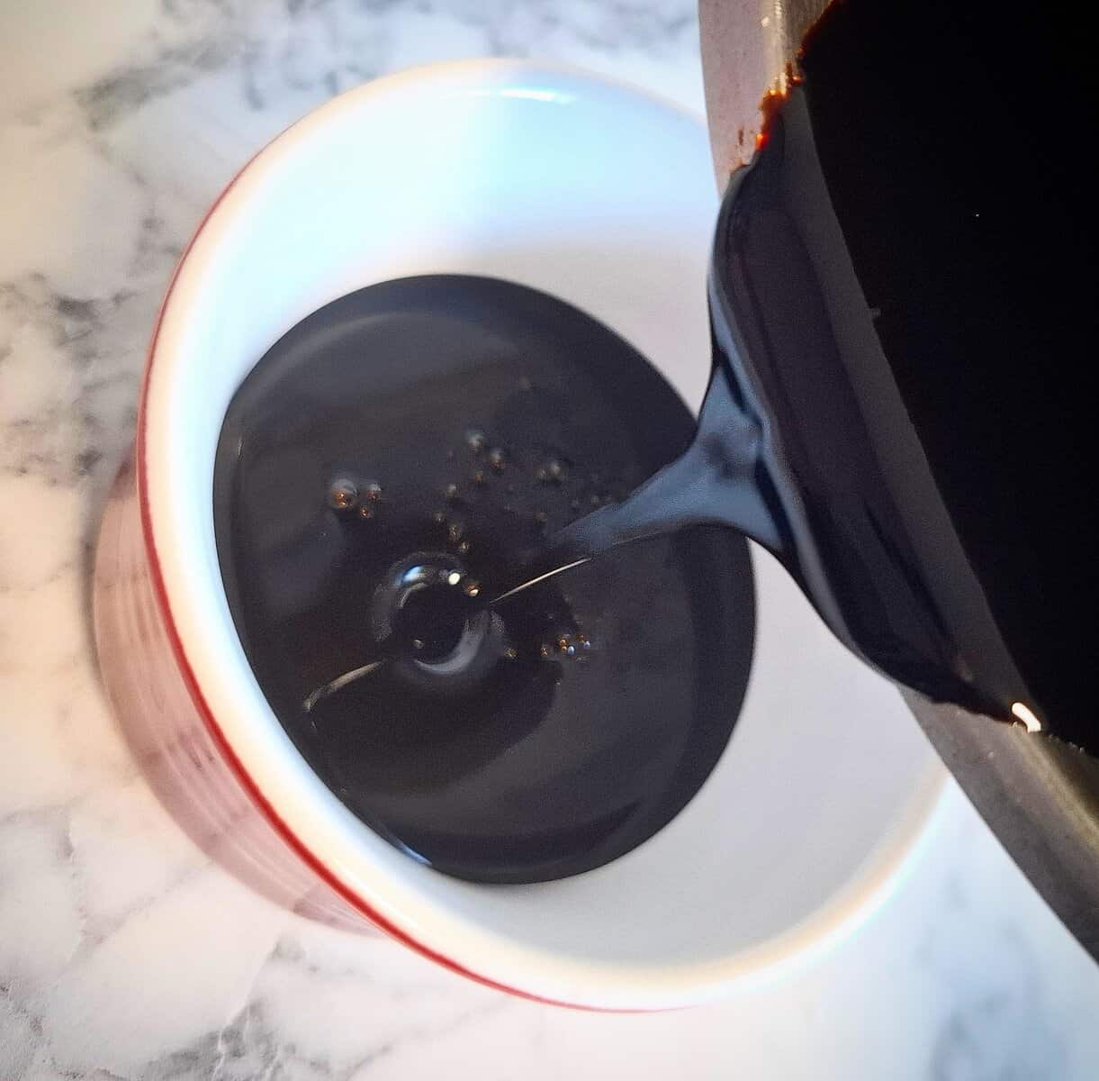 Pouring teriyaki sauce marinade from a saucepan into a white and red ramekin on a marbled bench top