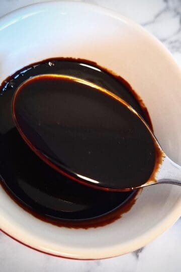 Teriyaki sauce marinade in a white and red ramekin on a marbled bench top. A spoon holding up some sauce in it.
