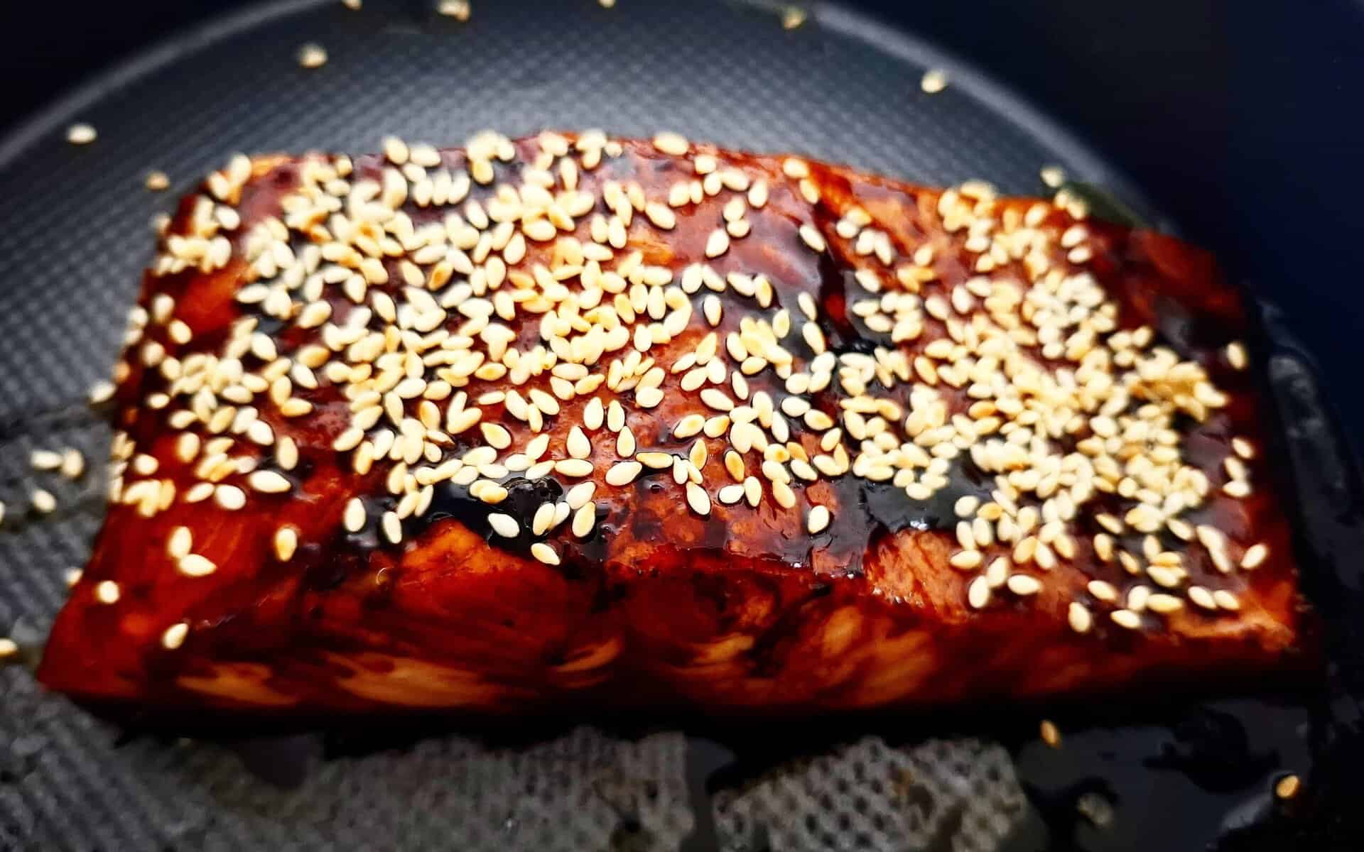 Teriyaki salmon topped with toasted white sesame seeds on a black oven tray