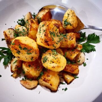 Mediterranean roasted potatoes served in a white bowl with parsley leaves garnish and a silver serving spoon placed in the bowl