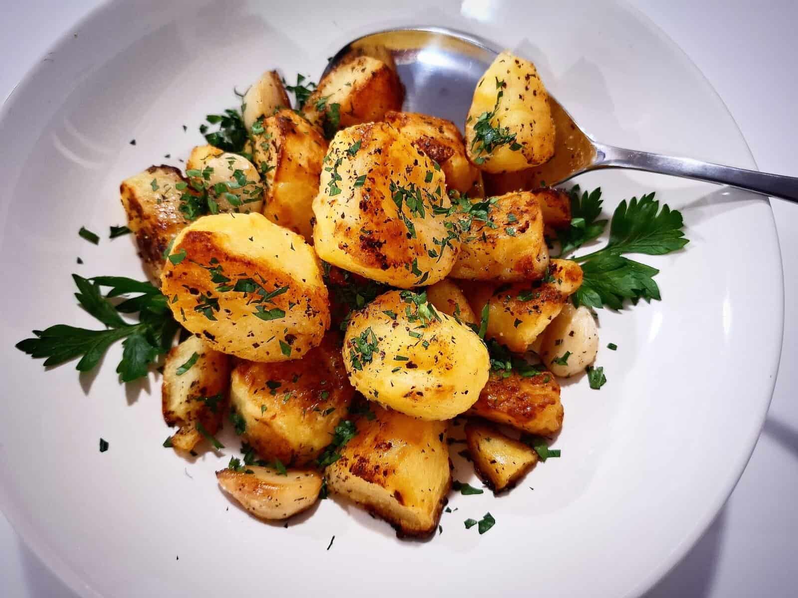 Mediterranean roasted potatoes served in a white bowl with parsley leaves garnish and a silver serving spoon placed in the bowl