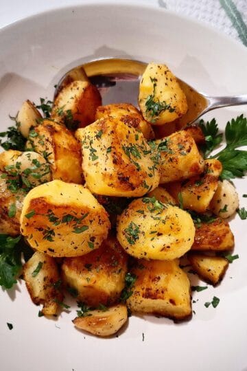 Mediterranean roasted potatoes served in a white bowl with parsley leaves garnish and a silver serving spoon placed in the bowl. A tea towel with green and white stripes next to it