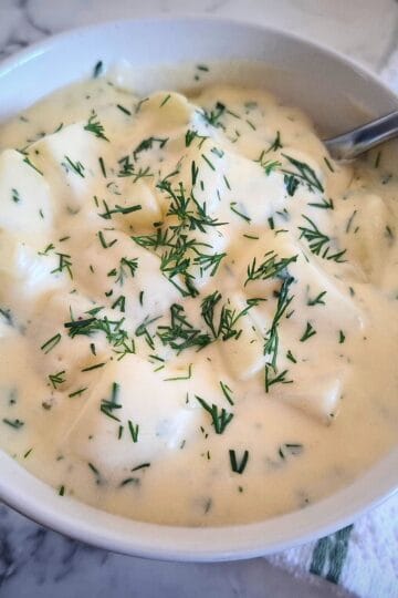 Dill creamed potatoes in a white bowl and a serving spoon in it. The bowl is placed on a marbled bench top with a white and green stripe towel next to it.