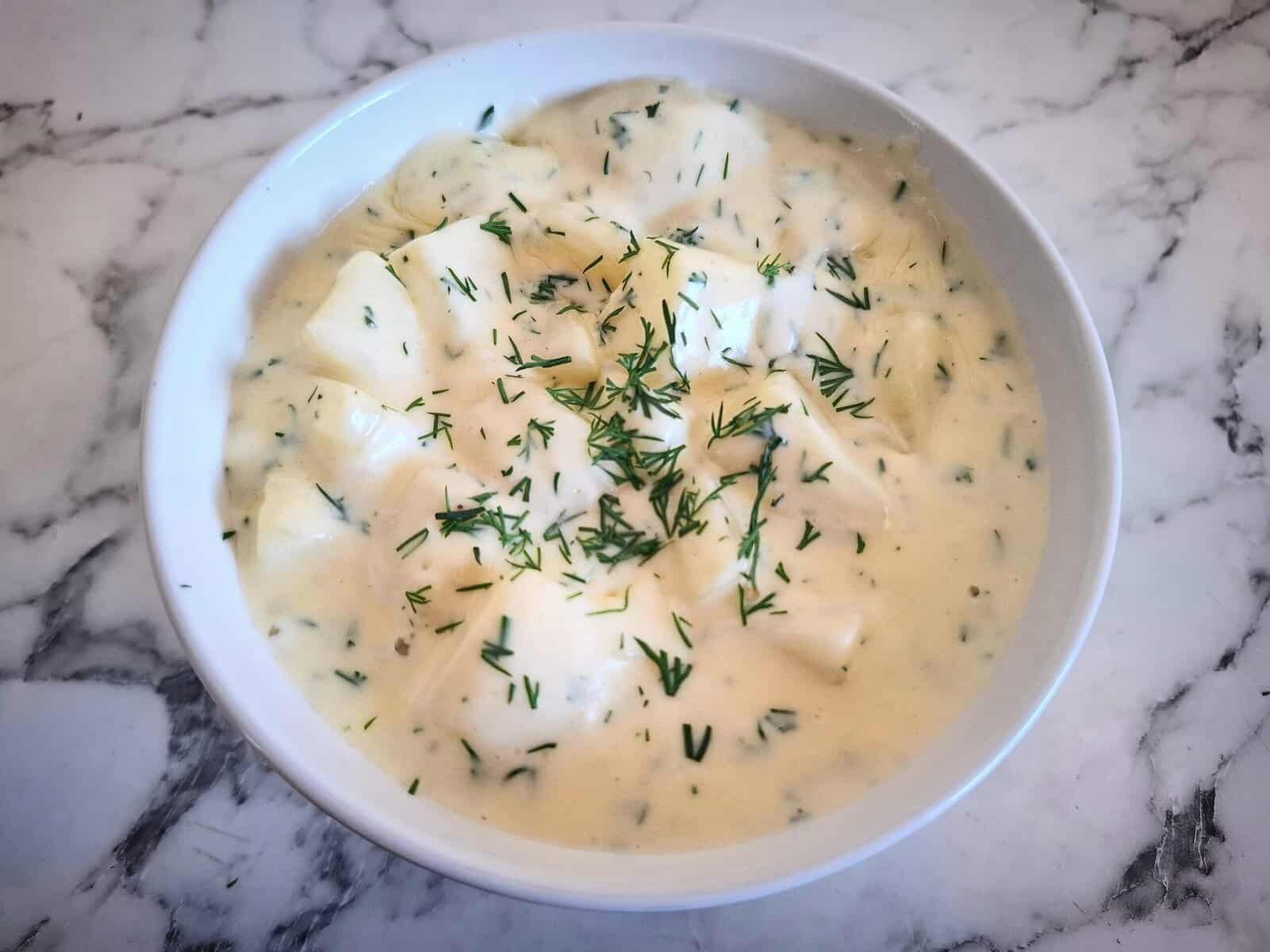 Dill creamed potatoes in a white bowl and a serving spoon in it. The bowl is placed on a marbled bench top with a white and green stripe towel next to it.