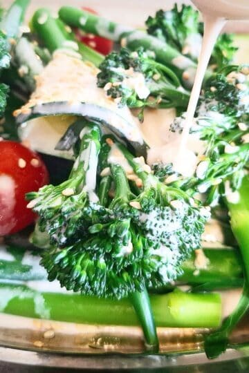 Steamed broccolini, green beans, zucchini slices and cherry tomatoes on an oval silver plate with tahini garlic dressing being drizzled over the top