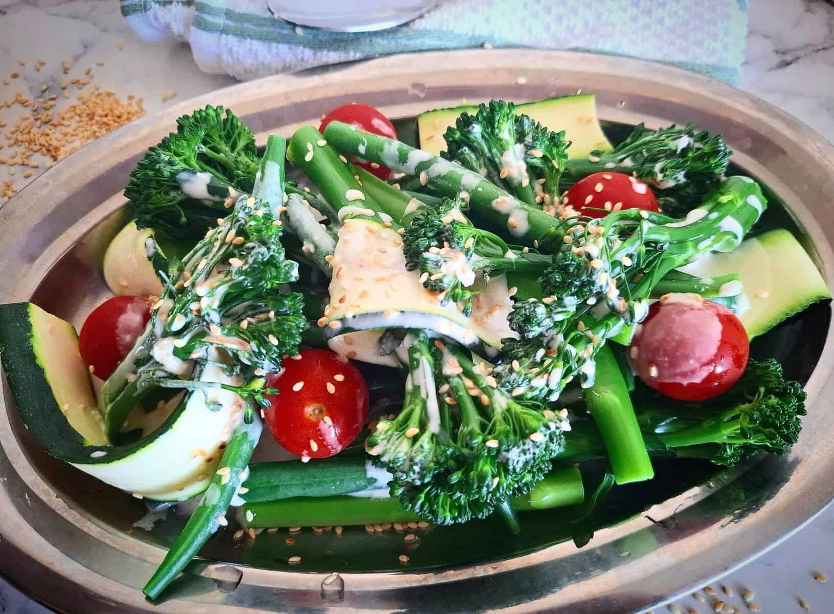 Steamed broccolini, green beans, zucchini slices and cherry tomatoes on an oval silver plate with tahini garlic dressing drizzled over the top garnished with sesame seeds. A white and green tea towel next to it. Sesame seeds sprinkled on the bench next to it.