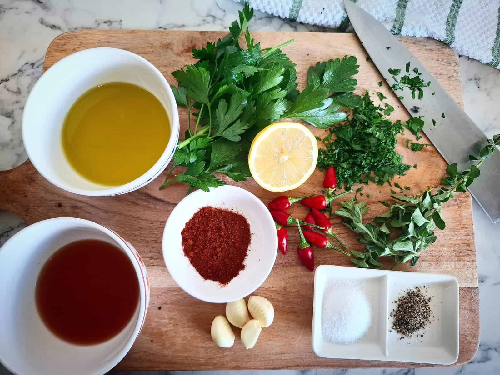 Ingredients for chimichurri sauce red on a wooden chopping board on a marbled bench top. A knife is placed in the top right corner of the chopping board with freshly cut parsley on it and chopped parsley next to it. A white and green striped tea towel is place above the chopping board.
