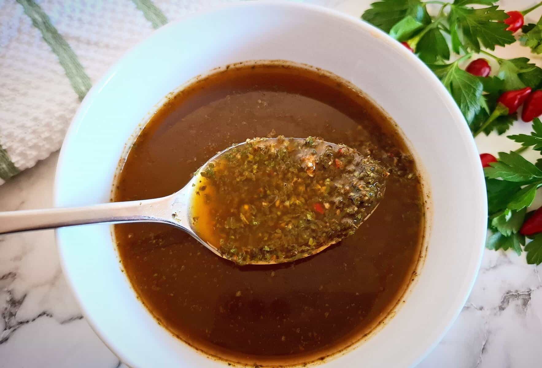Chimichurri sauce red in a white bowl on a marbled bench top with a spoon holding up sauce. Fresh parsley and chili next to it in the top right corner and a white and green striped tea towel in the top left corner next to the sauce.