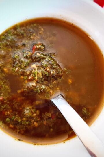 Red chimichurri sauce in a white bowl on a marbled bench top with a spoon in it. Fresh parsley and chili next to it in the top right corner and a white and green striped tea towel in the top left corner next to the sauce.