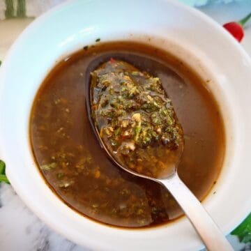Red chimichurri sauce in a white bowl on a marbled bench top with a spoon holding up sauce. Fresh parsley and chili next to it.