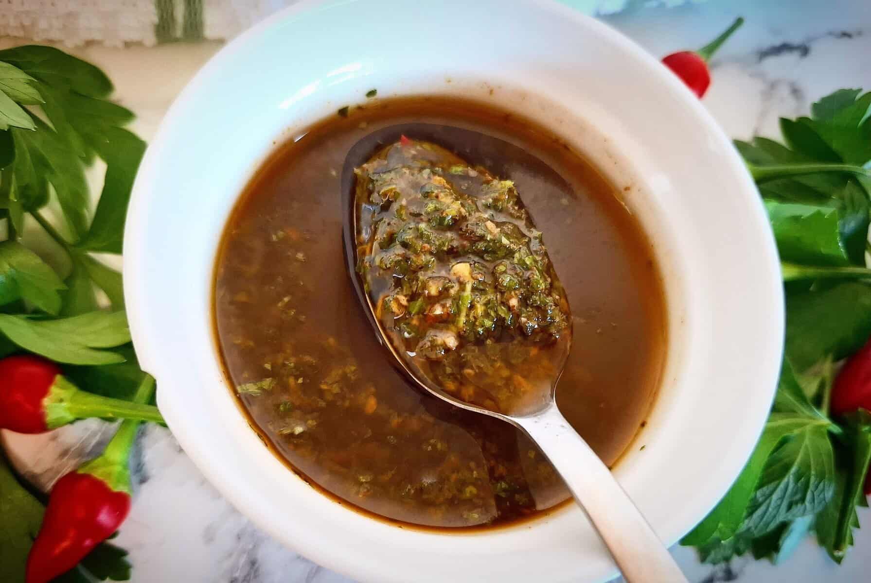Red chimichurri sauce in a white bowl on a marbled bench top with a spoon holding up sauce. Fresh parsley and chili next to it.