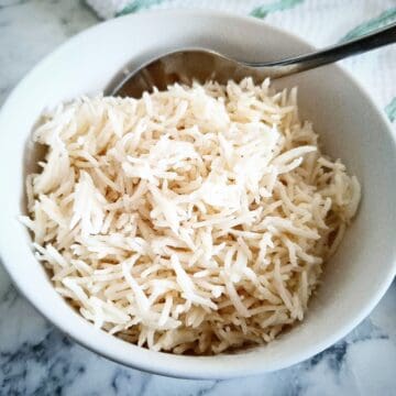 Coconut rice in a white serving bowl with a spoon in it on a marbled table and a white and green striped tea towel next to it in the top right corner.