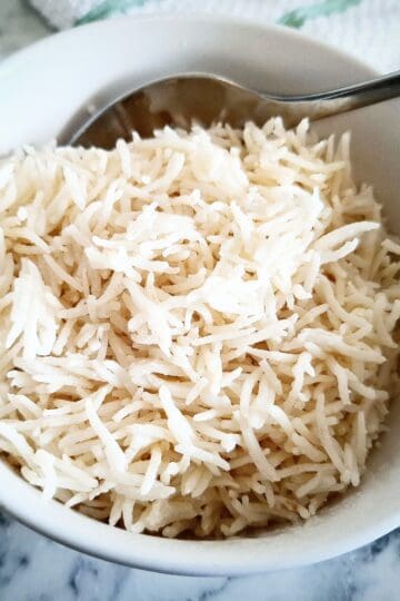 Coconut rice in a white serving bowl with a spoon in it on a marbled table and a white and green striped tea towel next to it in the top right corner.
