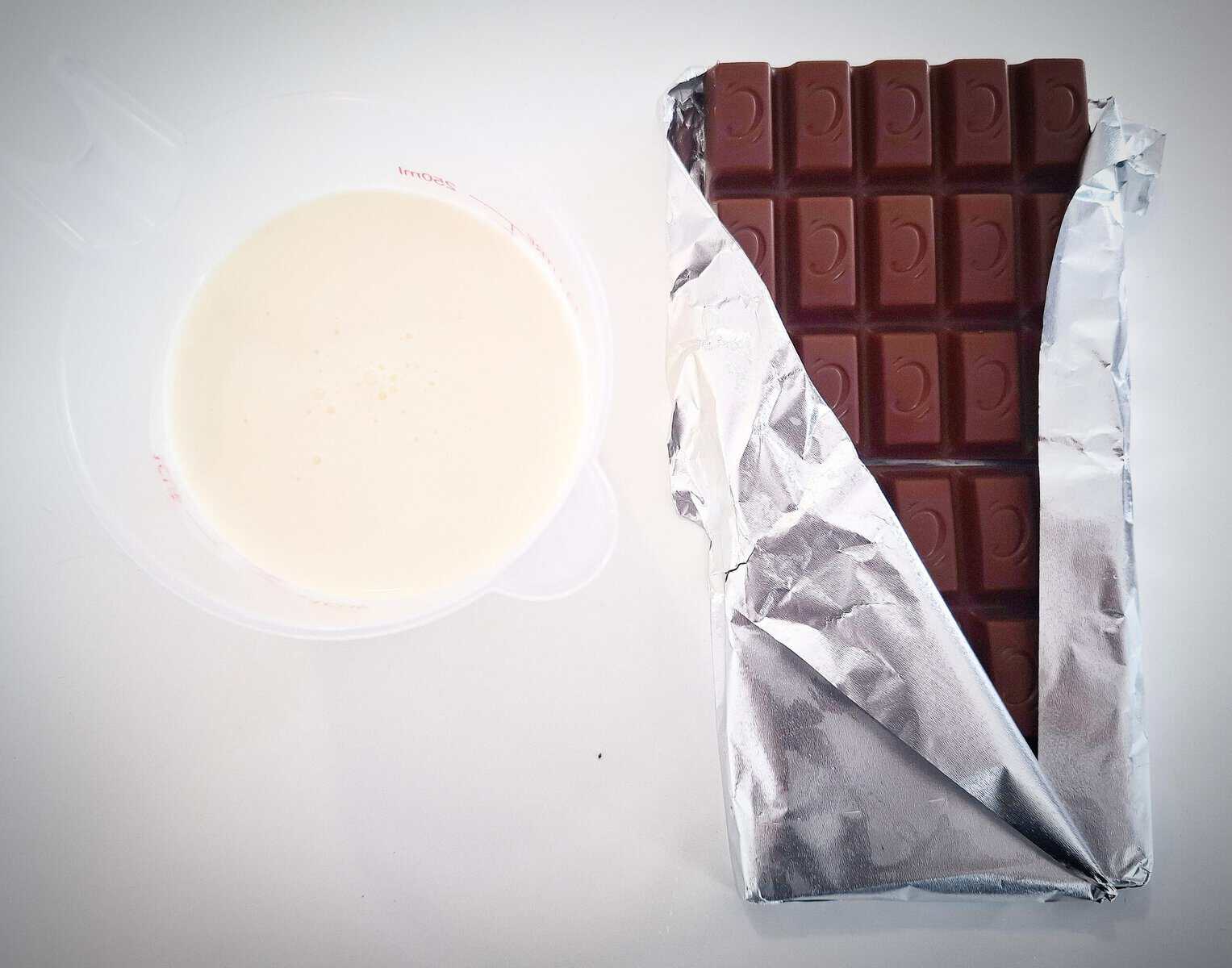 A white ramekin of cream and a chocolate bar in its foil wrapper on a white table top.