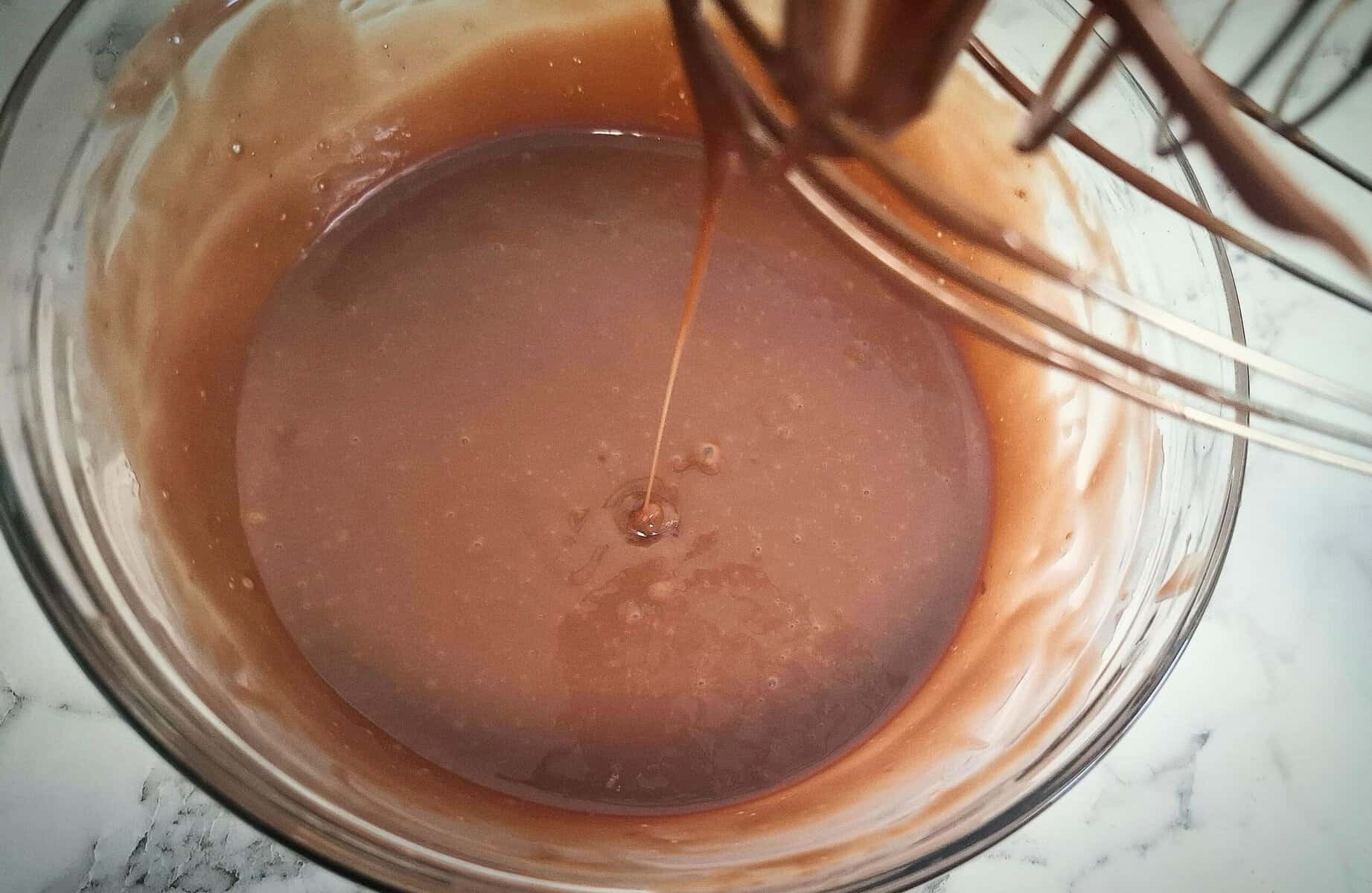 Melted ganache in a glass bowl with a whisk above it and chocolate dripping down from the whisk into the bowl
