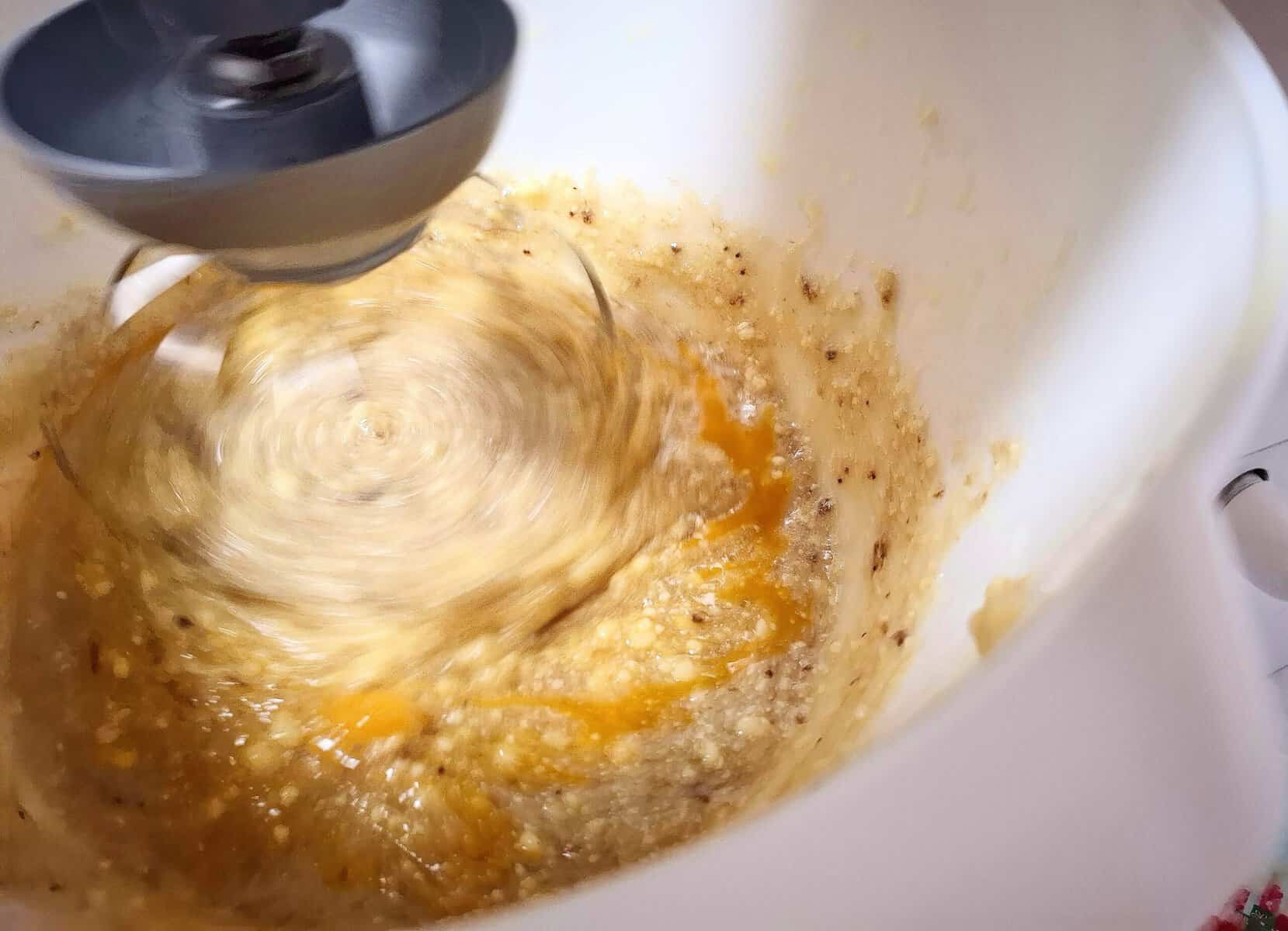 In a white kitchen mixer, mashed bananas and eggs are being added to the creamed butter and sugar mixture.
