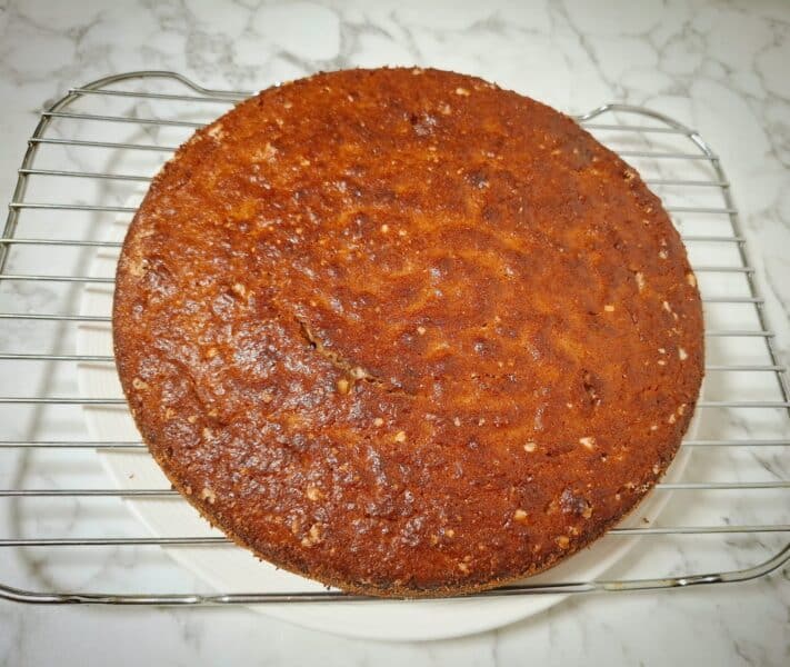 A banana chocolate cake resting on a wired wrack on top of a round white plate on a marbled bench top.