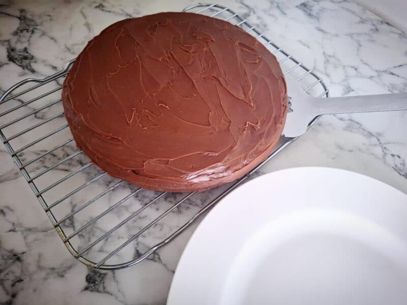 An iced banana chocolate cake on a wired wrack with a large spatula underneath the cake. A round white plate next to it.