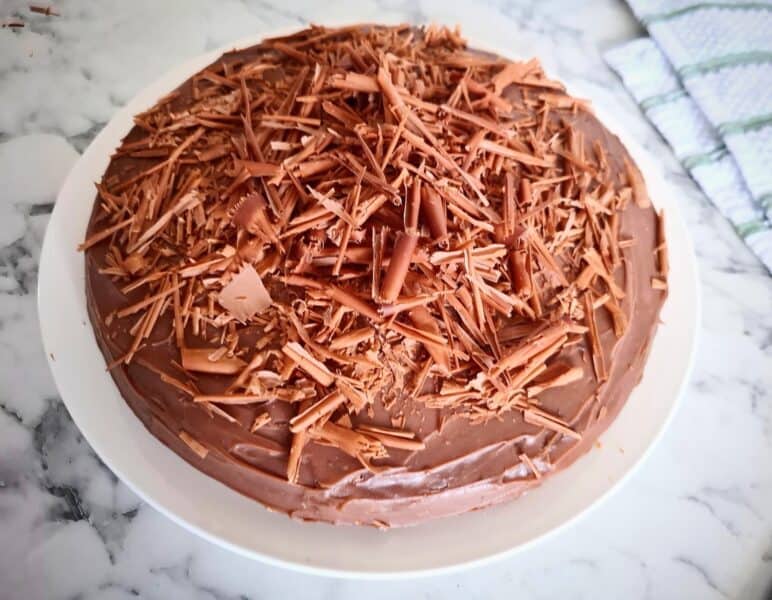 Iced banana chocolate cake decorated with chocolate shavings on top. The cake is served on a white round plate on a marbled bench top.