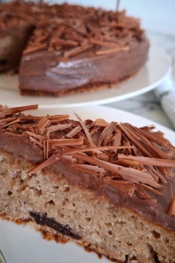 A portion of the iced and decorated chocolate banana cake is served on a white round plate and a fork upside down with it on the plate. The rest of the cake is on a separate plate in the back ground. A white and green striped tea towel with silver cake serving cutlery on top next to it.