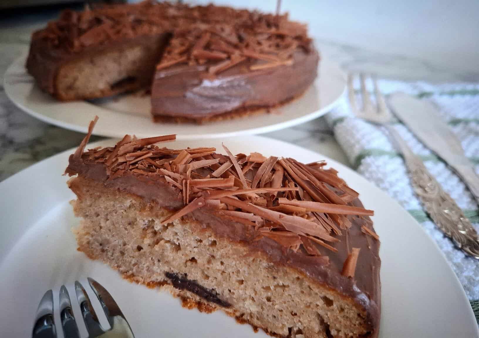 A portion of the iced and decorated chocolate banana cake is served on a white round plate and a fork upside down with it on the plate. The rest of the cake is on a separate plate in the back ground. A white and green striped tea towel with silver cake serving cutlery on top next to it.